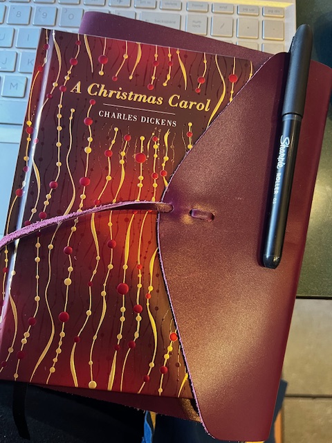 Picture of book, A Christmas Carol by Charles Dickens, resting inside a maroon leather journal, with black pen, all atop a silver laptop keyboard. 