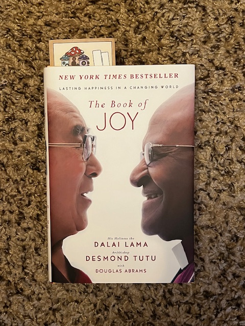 Cover of The Book of Joy by Dalai Lama and Desmond Tutu, on brown carpet with mushroom bookmark. 
