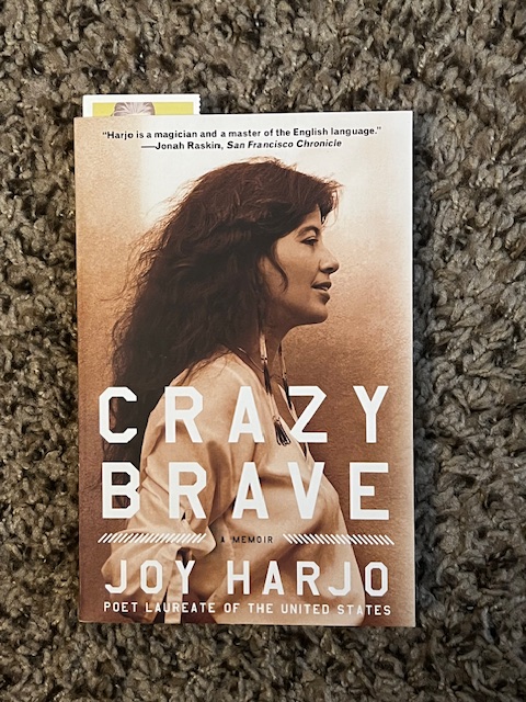 Paperback book--Crazy Brave by Joy Harjo--with bookmark peeking out, sitting atop tan fabric background. 